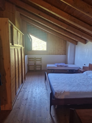 A room with a sloped ceiling, wooden flooring, a bed, a chair, a small table, and a window with a view of trees.