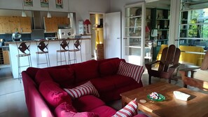A living room with a red sofa, wooden chairs, a wooden table, and a kitchen in the background. - Cap Ferret maison familiale, plage à 5 mn et proche des commerces. (Lège-Cap-Ferret)