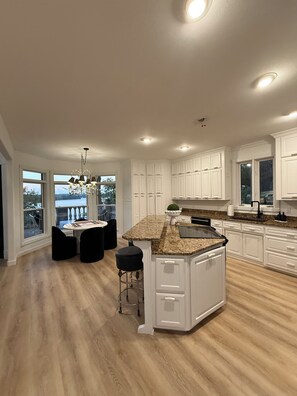 A spacious kitchen with white cabinetry, a granite countertop island, and a view through large windows. - Gorgeous Lakefront 5 bedroom with Stunning Views (Rogers)