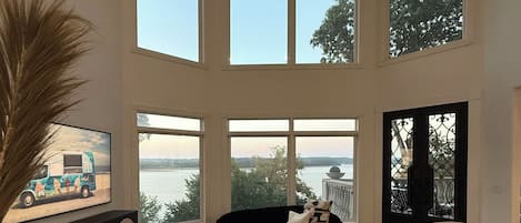 A spacious living room with a large window overlooking a body of water, a black sofa, a coffee table, and a television on a stand.