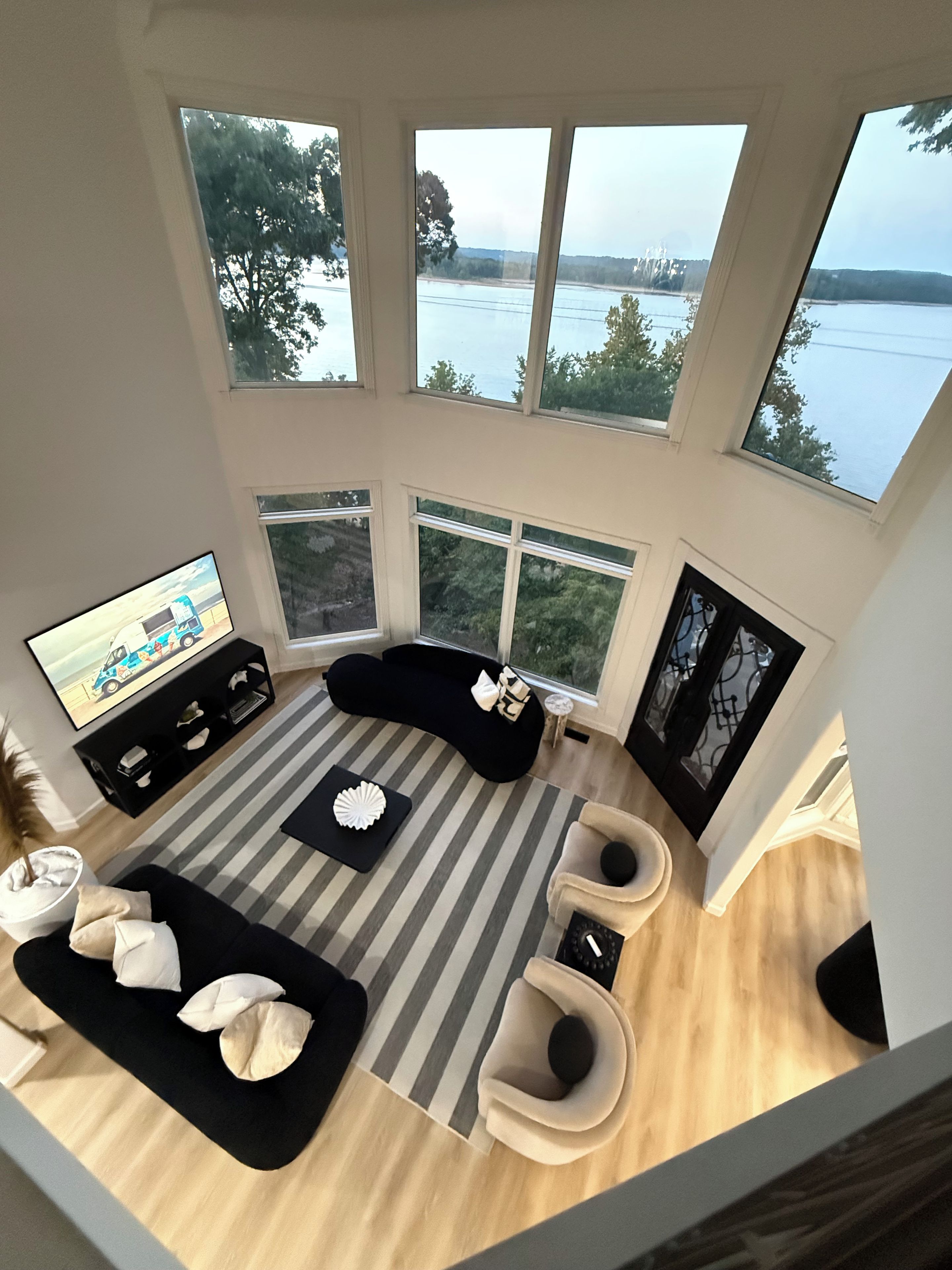 A modern living room with a large window overlooking a body of water, a black sofa, a striped rug, and a television on a stand.