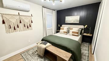 Une chambre à coucher moderne comprenant un lit, des tables de chevet, un banc, un climatiseur fixé au mur, un luminaire au plafond et une fenêtre avec des rideaux.