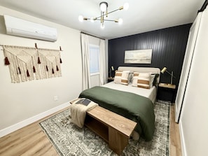 A modern bedroom with a bed, bedside tables, a bench, a wall-mounted air conditioner, a ceiling light fixture, and a window with curtains.