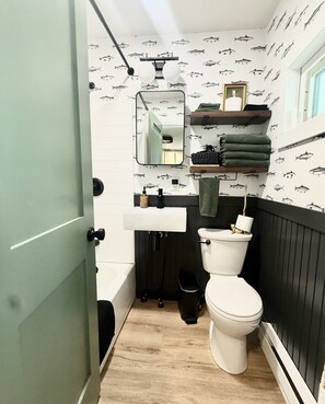 A compact bathroom with a toilet, sink, and mirror. There are shelves above the sink with towels and toiletries. The walls have a fish pattern.