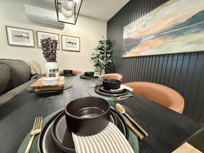 A dining area with a dark wooden table set for two, a sofa, a plant, and framed artwork on the walls.