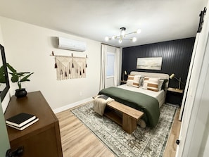 Une chambre à coucher comprenant un lit, une table de chevet, un banc, un appareil de climatisation, des éléments de décoration murale et une fenêtre avec des rideaux.