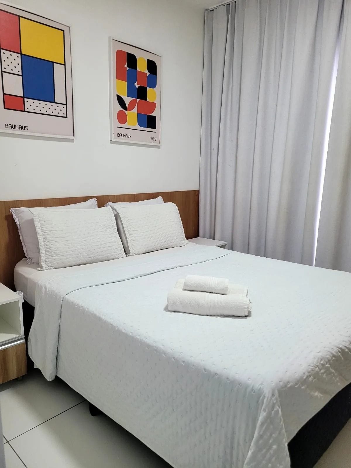 Una cama bien hecha con ropa blanca y dos almohadas, una mesita de noche con una lámpara y dos obras de arte abstracto en la pared.
