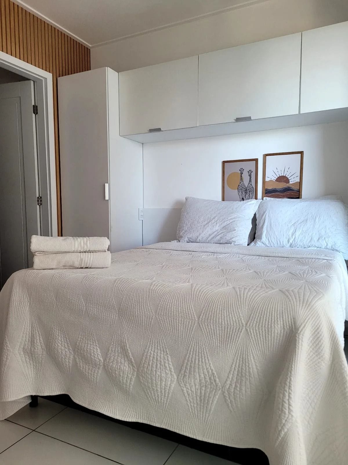 Quarto com cama, roupa de cama branca, um guarda-roupa e duas imagens emolduradas na parede.