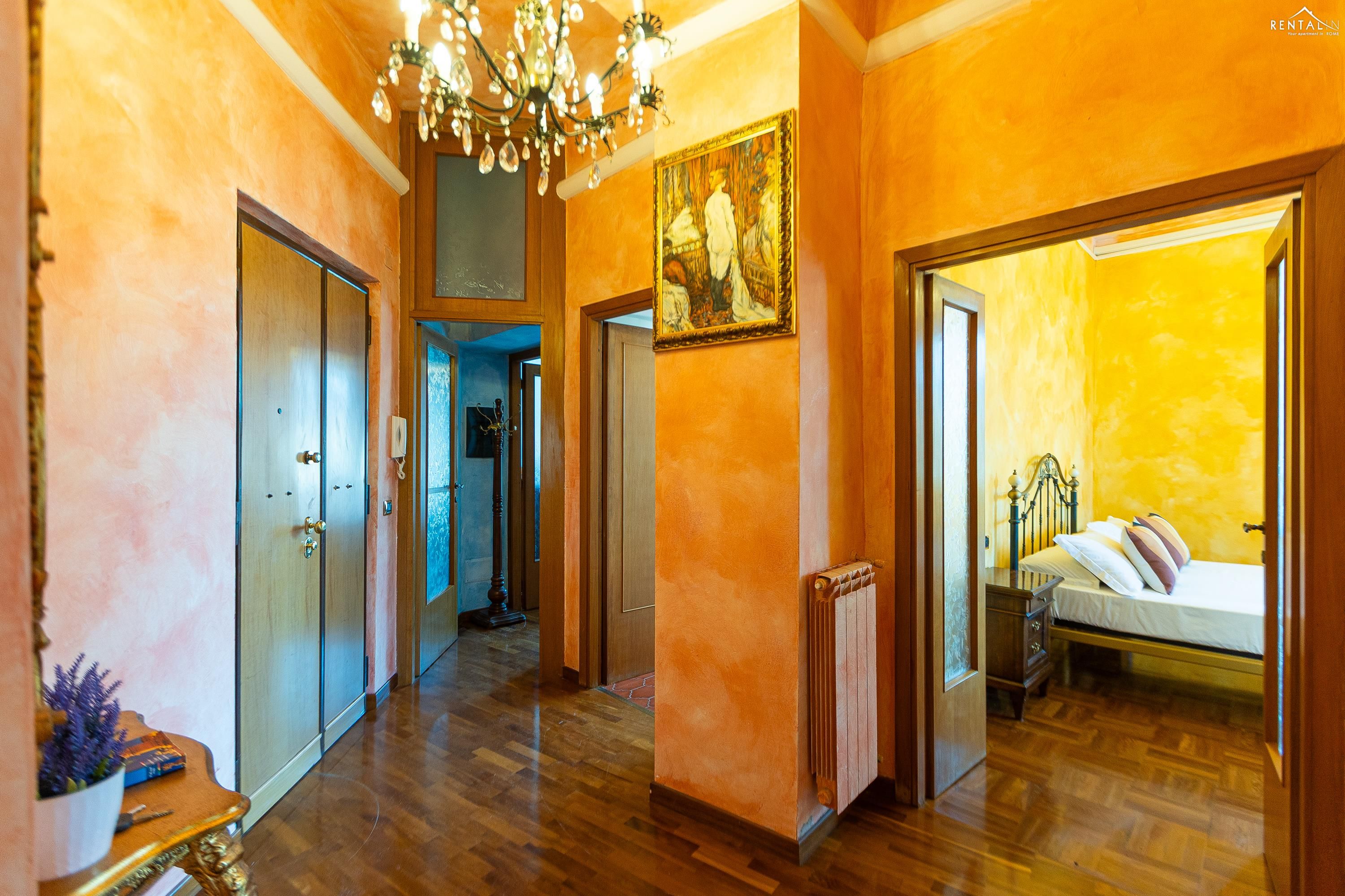 A hallway with wooden floors, a chandelier, a painting, and a bed in the adjacent room.