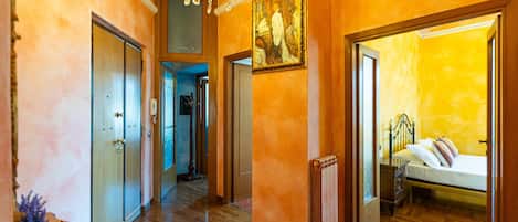 A hallway with wooden floors, a chandelier, a painting, and a bed in the adjacent room.