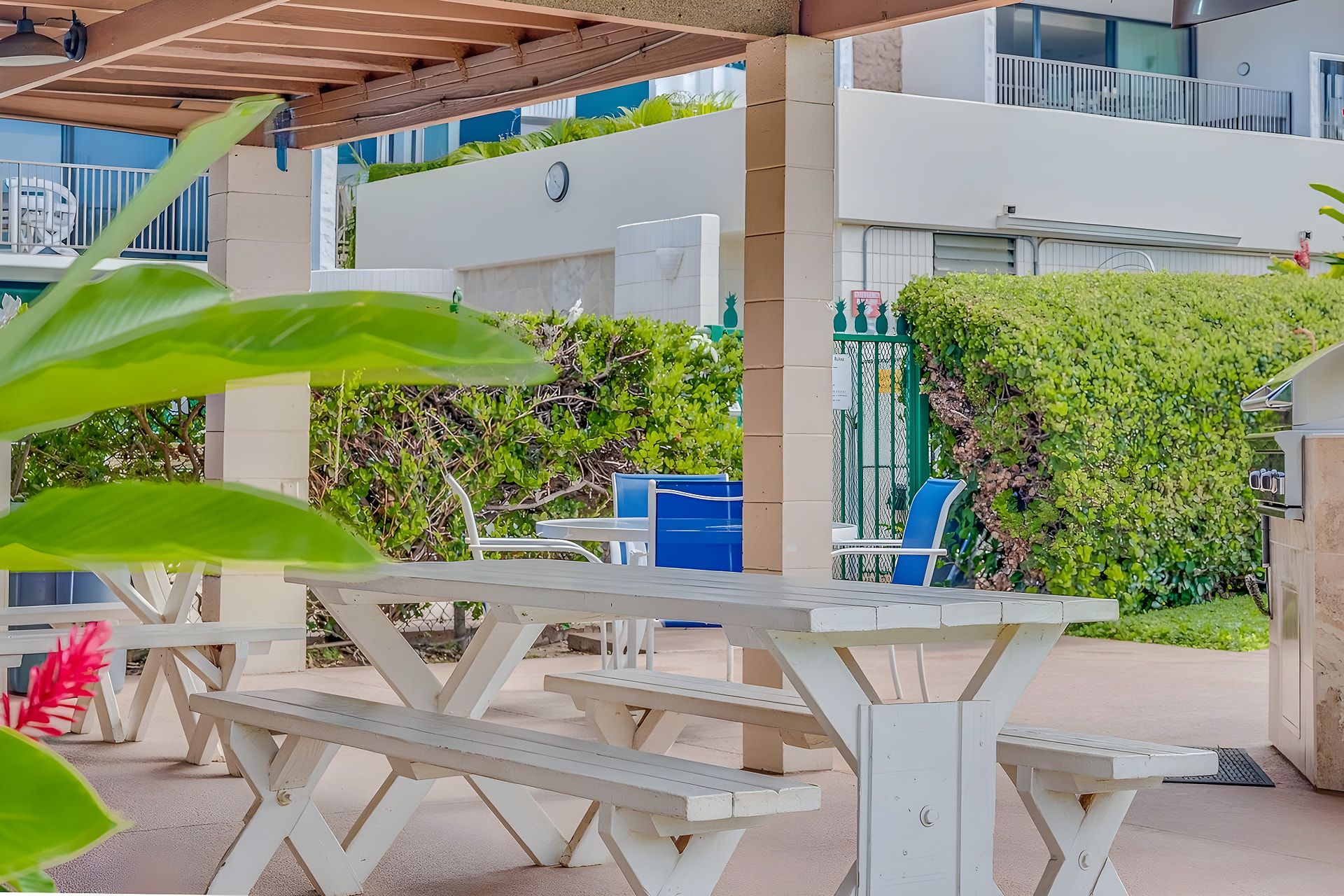 Un espacio al aire libre techado con mesas de picnic, un edificio blanco y setos verdes.