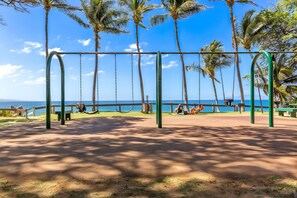 A swing set with a view of the ocean, surrounded by palm trees and a clear sky. - Kihei, Maui Parkshore, Spacious 4 BR, Sleeps 12 (Kihei)