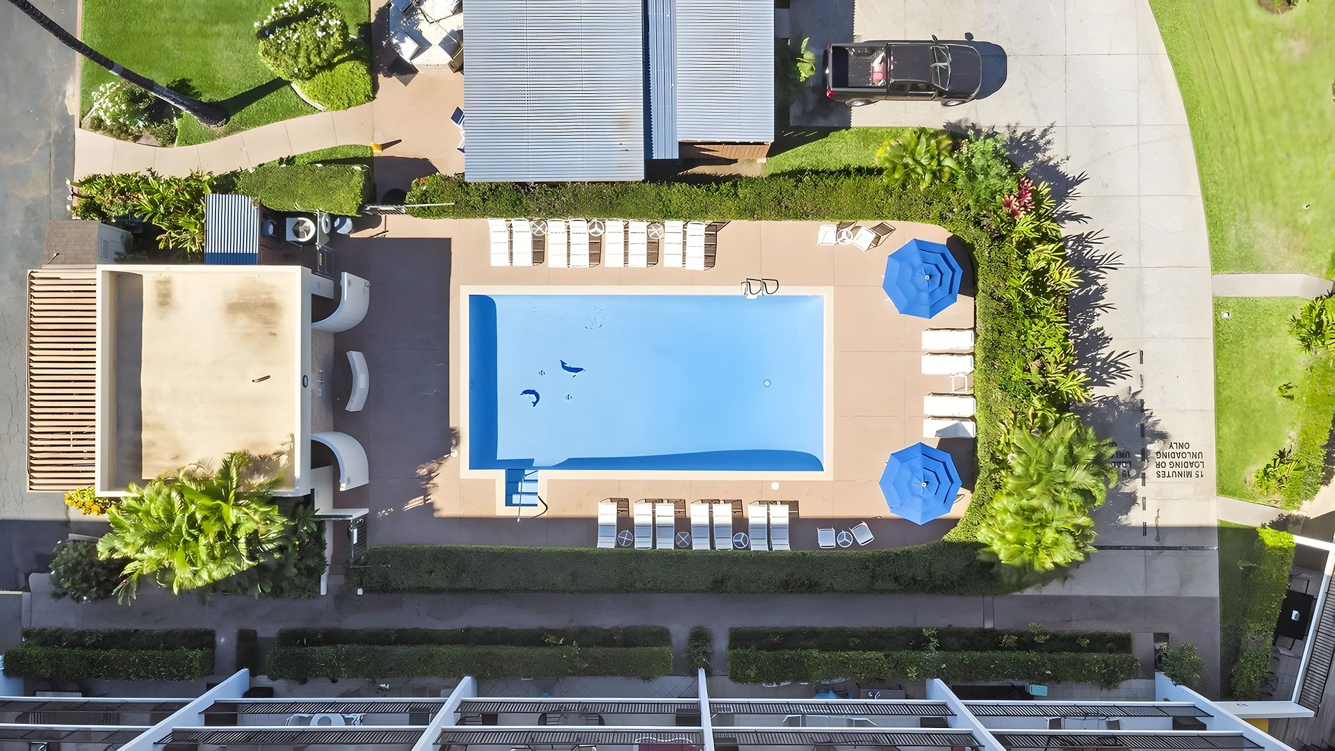 Vista aérea de una propiedad con piscina, sillas de descanso, sombrillas y un estacionamiento.