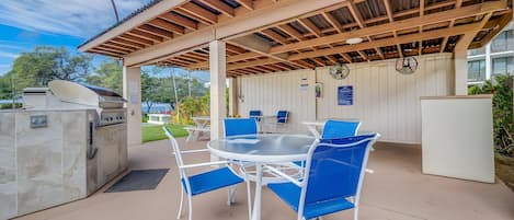 Eine Außenpatio mit einem runden Tisch, blauen Stühlen, einem Grill und einem überdachten Bereich mit Holzboden.