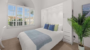 A bedroom with a bed, a window with blinds, a bedside table, a plant, and a painting on the wall. - Walk to the Beach from a 4-Bedroom Villa with Bikes and Beach Gear Included (Marco Island)