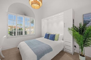 Un dormitorio con cama, ventana con persianas, mesita de noche, una planta y un cuadro en la pared.
