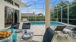 An outdoor pool area with a covered seating area, a dining table set with plates and glasses, and a view of a residential building in the background. - Walk to the Beach from a 4-Bedroom Villa with Bikes and Beach Gear Included (Marco Island)