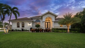 A two-story house with a large front lawn, a well-maintained garden, and a clear sky. - Walk to the Beach from a 4-Bedroom Villa with Bikes and Beach Gear Included (Marco Island)