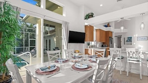 A modern dining area with a large table set for six, surrounded by white chairs. - Walk to the Beach from a 4-Bedroom Villa with Bikes and Beach Gear Included (Marco Island)