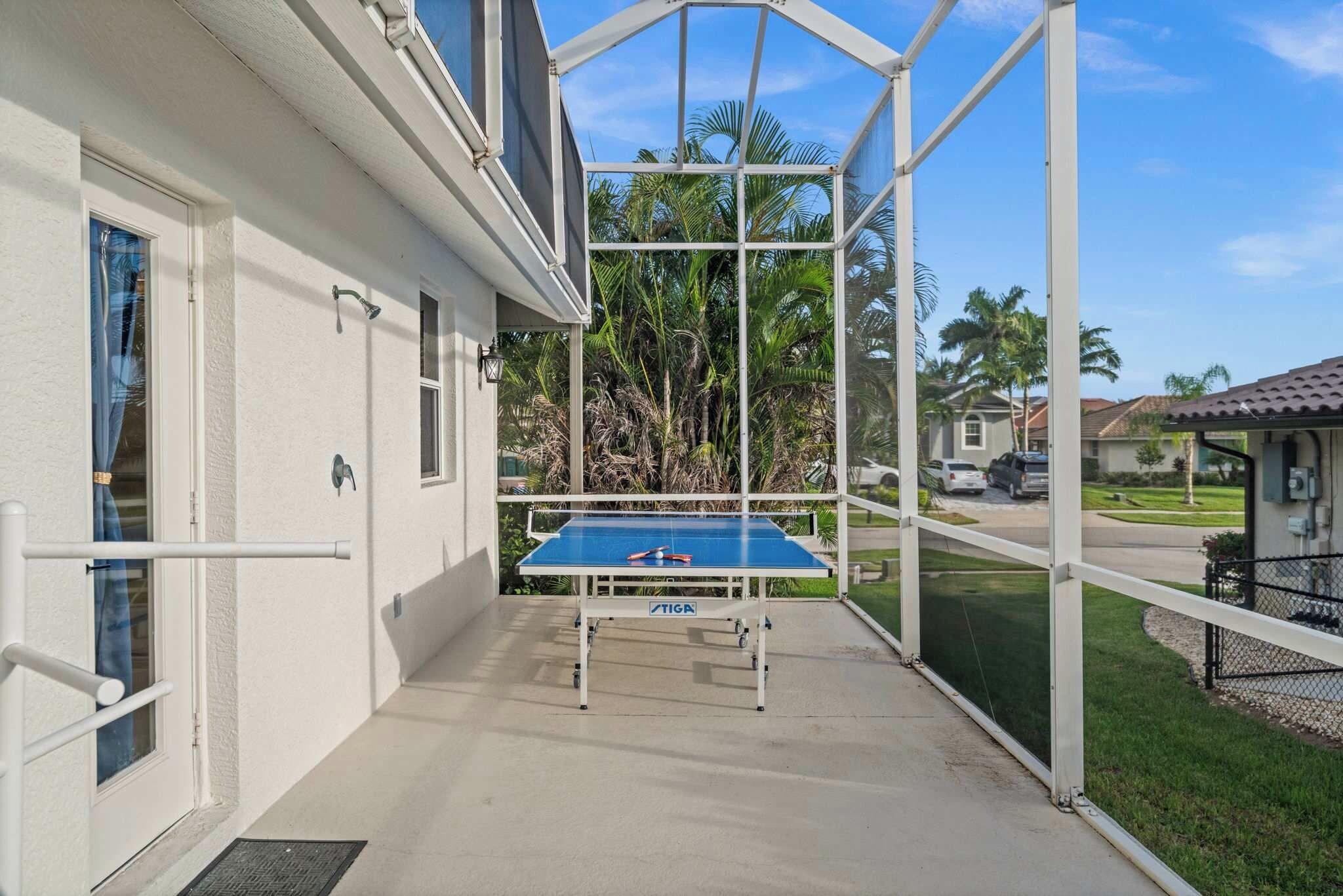 A covered outdoor area with a blue table tennis table, a white building, and a view of palm trees and houses.