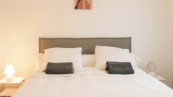 Una cama bien hecha con ropa blanca y dos almohadillas oscuras, una lámpara de noche y un cuadro en la pared.
