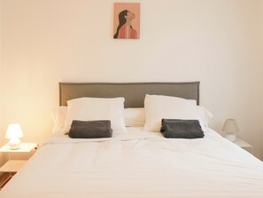Una cama bien hecha con ropa blanca y dos almohadillas oscuras, una lámpara de noche y un cuadro en la pared.