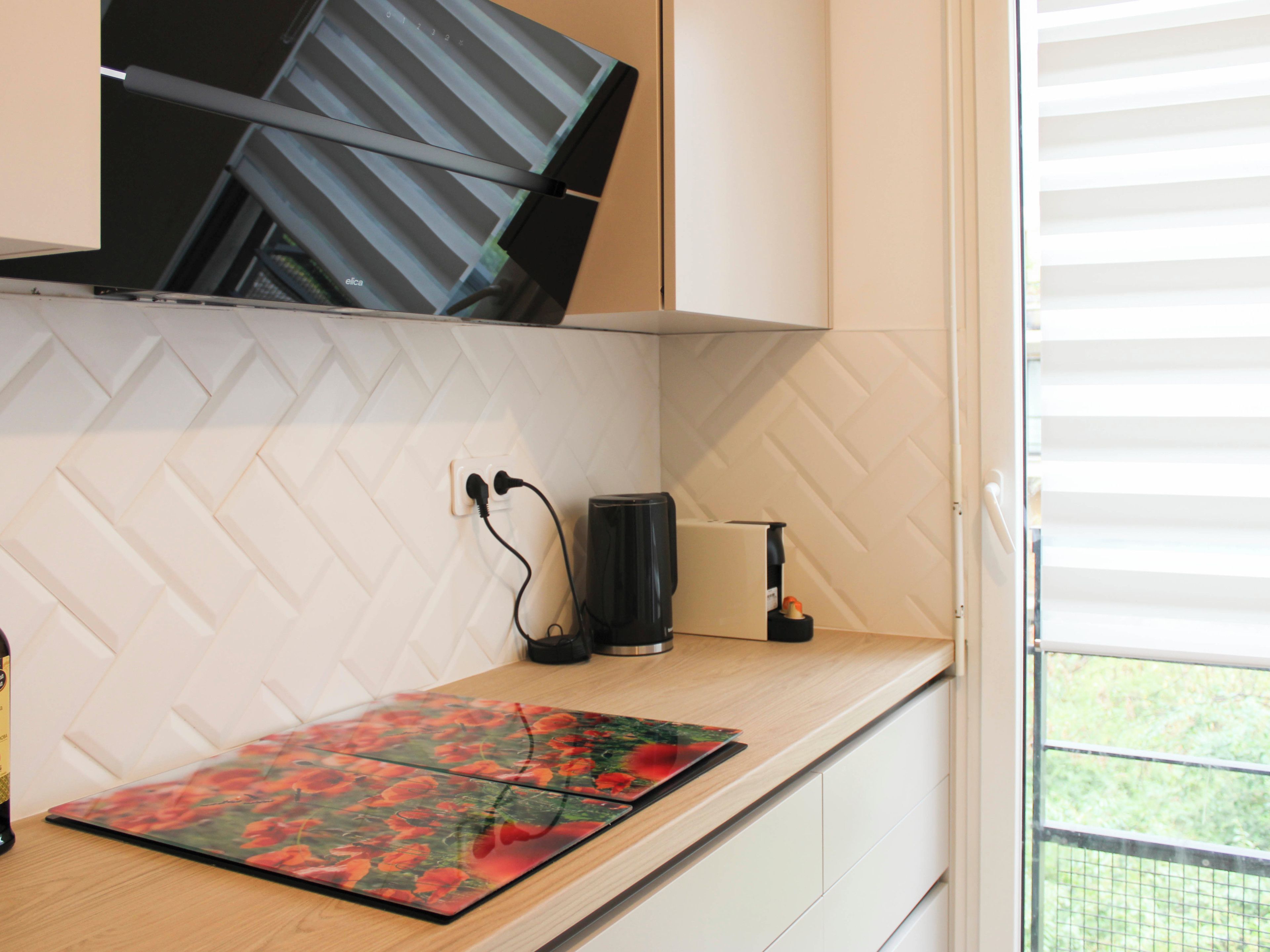Une cuisine avec un dosseret de carreaux blancs disposés en motif chevron, un plan de travail en bois, un set de table à motifs floraux rouges, une machine à café noire et une prise électrique.