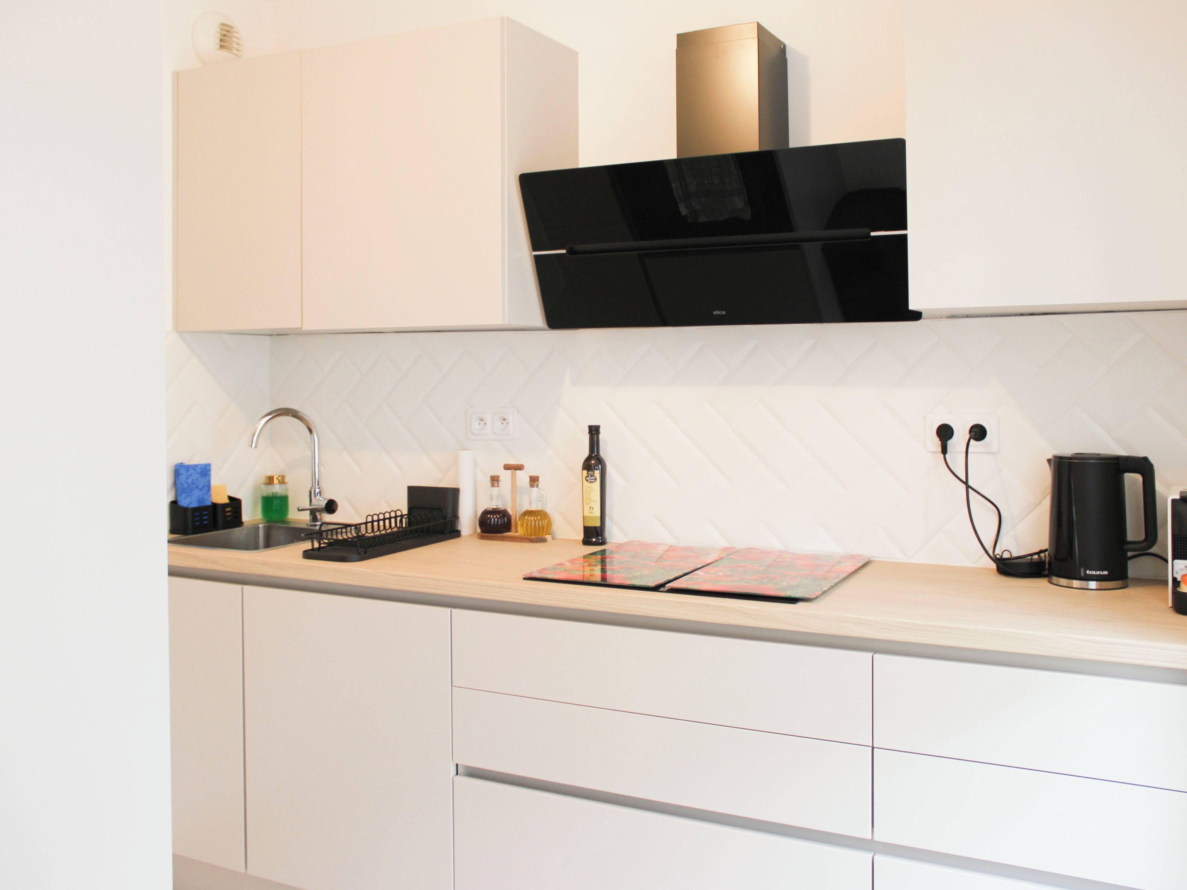 A modern kitchen with white cabinetry, a black range hood, a sink with a faucet, and a kettle on the countertop.