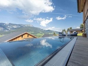 A clear sky with a mountainous landscape in the background, a swimming pool with a wooden deck, and a wooden building with a sloped roof. - One-bedroom Cabin (Haus)