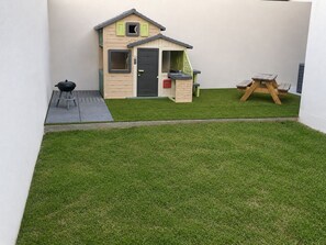 Ein kleines Spielhaus mit Tür und Fenstern, ein Grill und eine Holzschaukel im Garten.