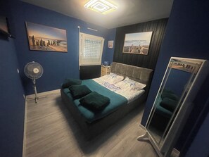 Ein Schlafzimmer mit einem Bett, einem Standventilator, einem Fenster mit Jalousien und zwei Wandbildern.