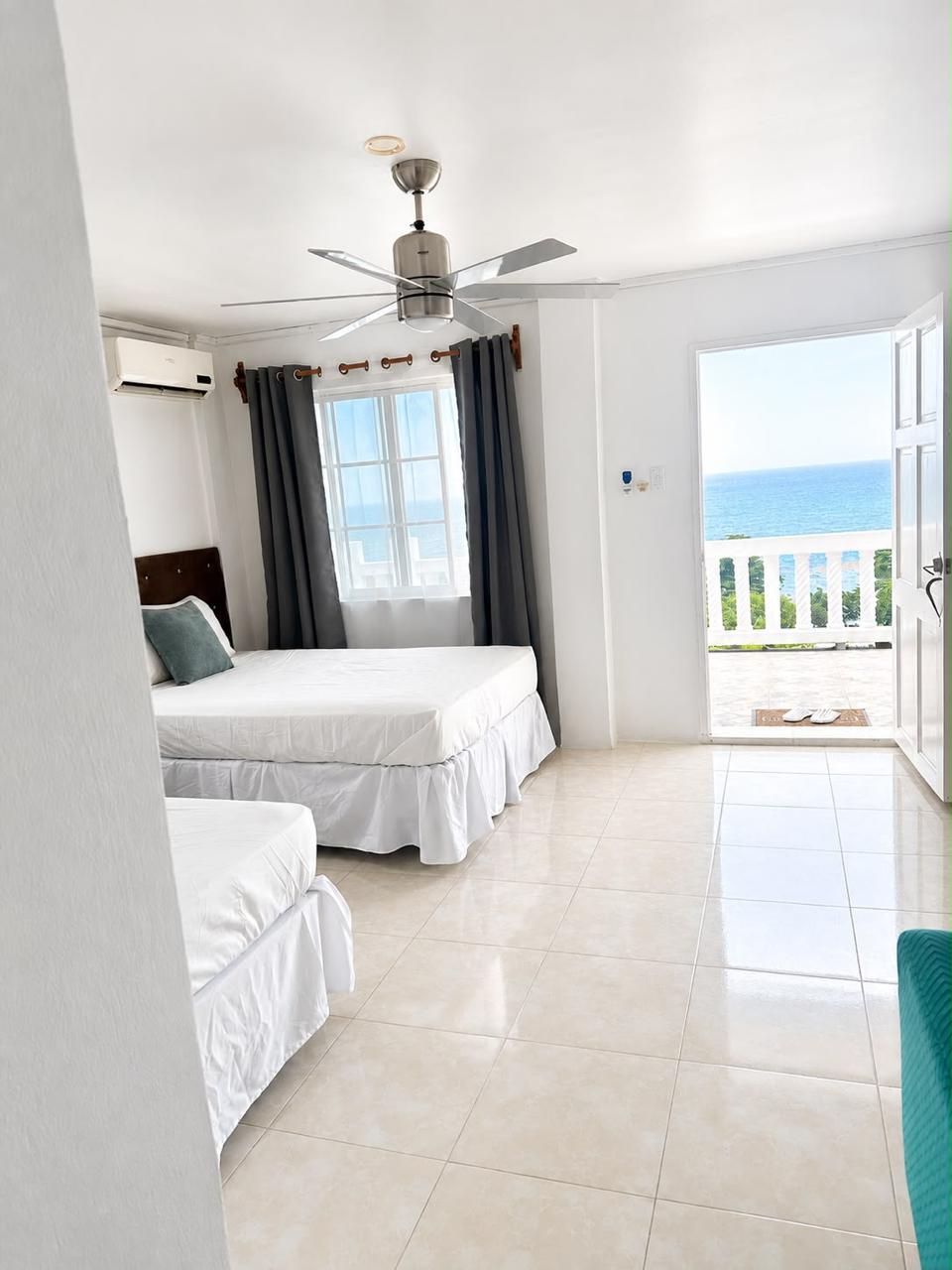 A hotel room with two beds, a ceiling fan, an air conditioning unit, a window with curtains, and a balcony with a view of the sea.