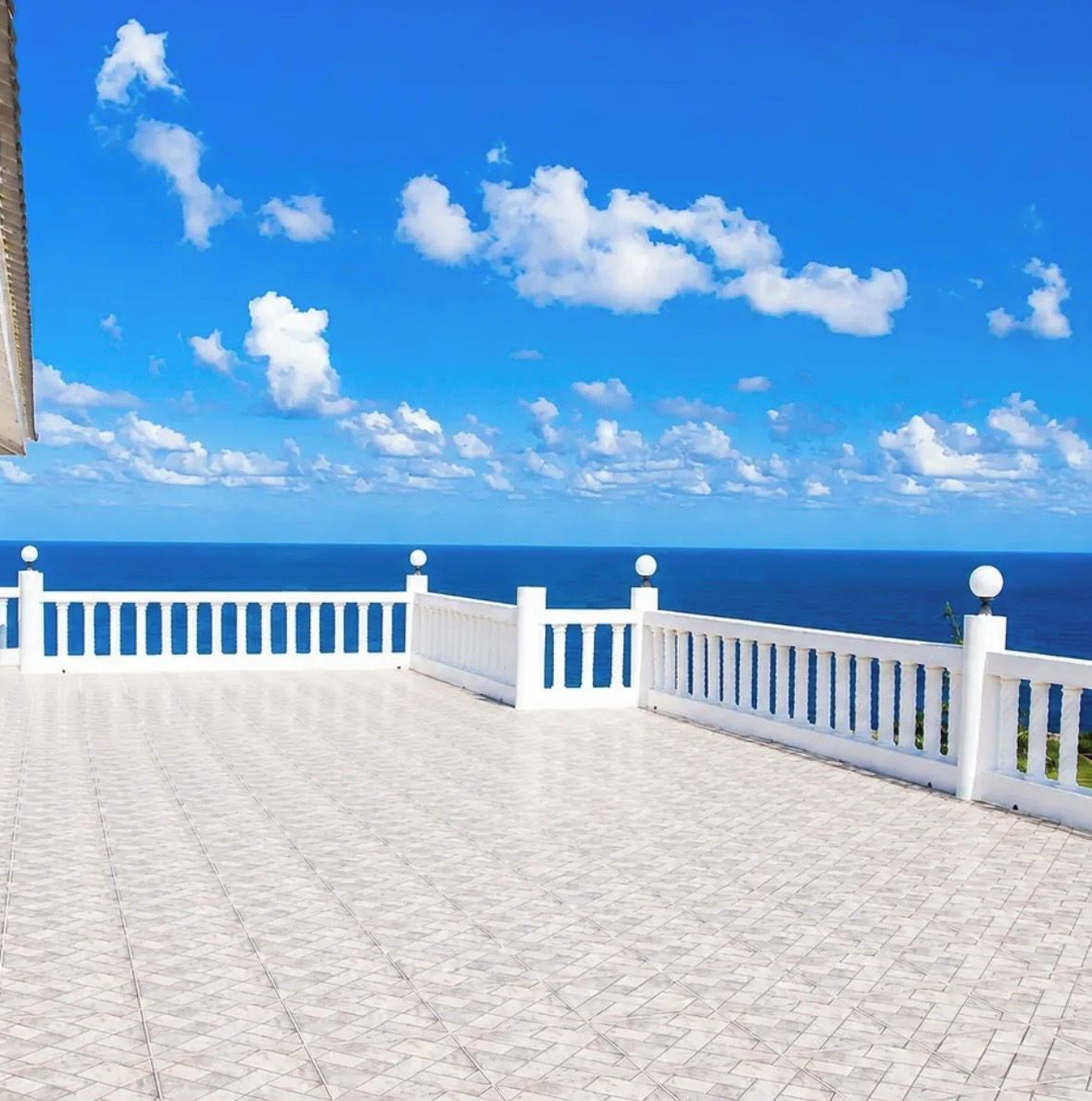 A white balcony with a tiled floor and a white railing, overlooking a clear blue sky and ocean.