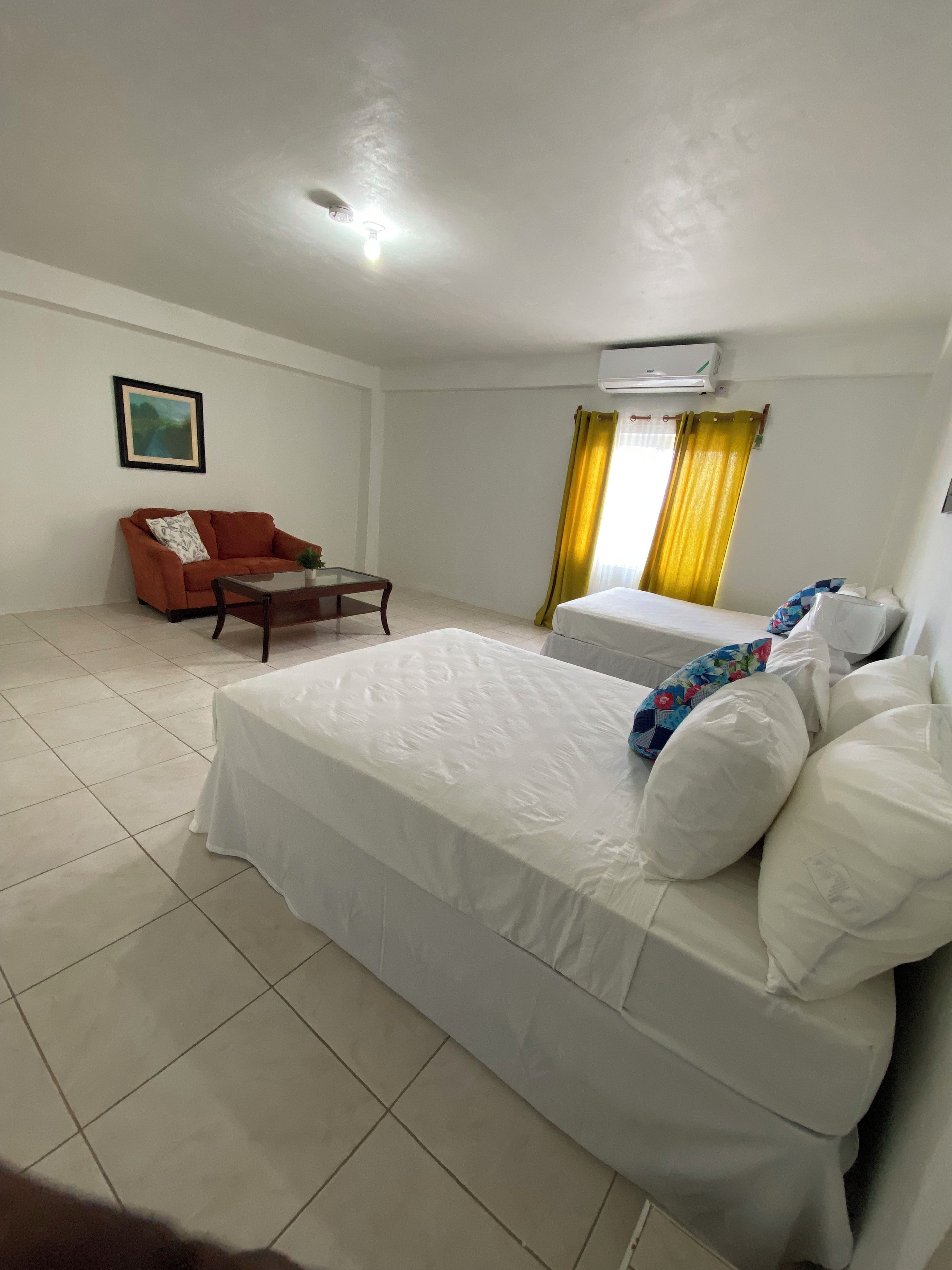 A hotel room with a bed, a sofa, a small table, and an air conditioning unit.