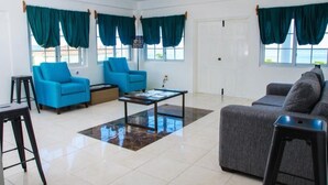 Ein Wohnzimmer mit zwei blauen Sesseln, einem grauen Sofa, einem Couchtisch und Meerblick durch große Fenster mit türkisfarbenen Vorhängen.