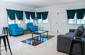 A living room with two blue armchairs, a grey sofa, a coffee table, and a view of the sea through large windows with teal curtains. - Da Villa Juanita (Hordley)
