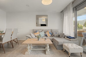 Una sala moderna con un sofá, una mesa de centro y un reposapiés, un comedor con sillas y un amplio ventanal con vistas.