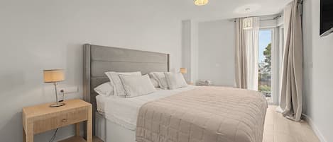 A bedroom with a bed, bedside tables, a nightstand, a lamp, curtains, and a view of the outdoors.