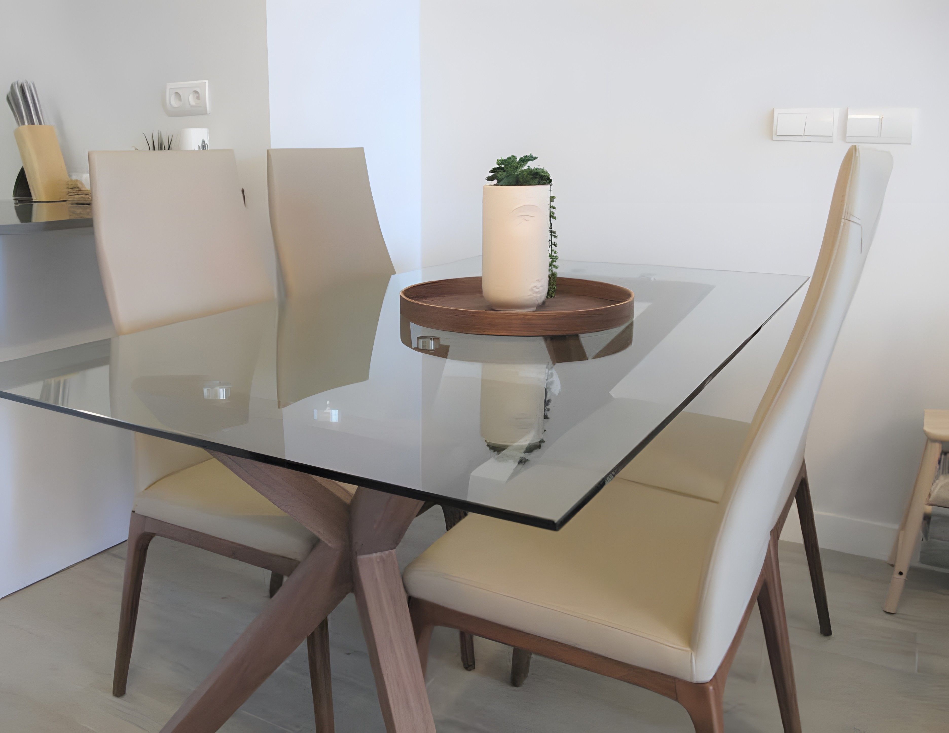 Un comedor moderno con una mesa de vidrio, dos sillas, una planta en maceta y una mesita de madera.