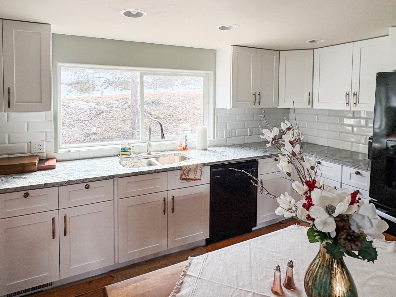 A kitchen with white cabinets, a black dishwasher, and a window with a view of a snowy landscape.
