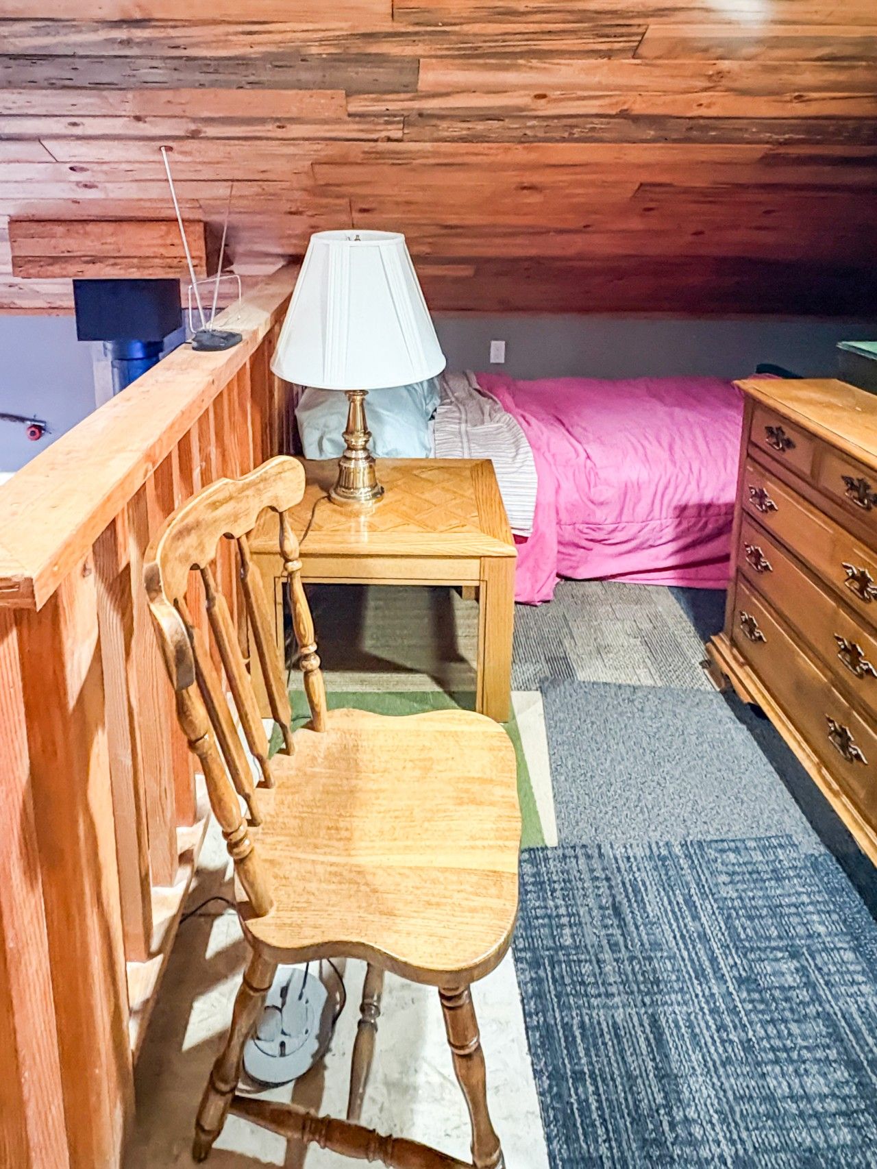 Ein Holzsessel, ein kleiner Tisch mit einer Lampe, ein Bett mit einer rosa Decke und eine Kommode in einem Raum mit Holzwänden und einer geneigten Decke.