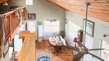 A spacious loft with a high ceiling, wooden beams, a dining area with a table and chairs, a blue armchair, and a checkered sofa.