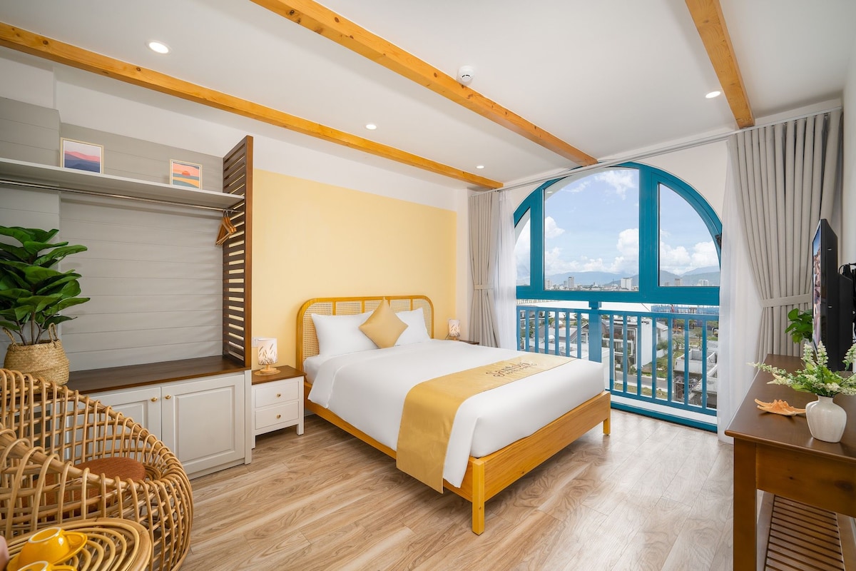 Quarto de hotel com cama, mesa de cabeceira, cadeira de vime, varanda e vista, e janela com cortinas.