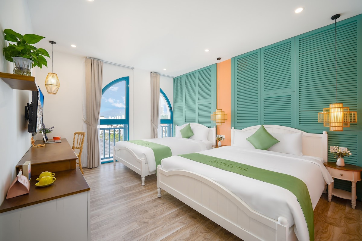 Quarto de hotel com duas camas, uma escrivaninha, uma cadeira, varanda e vista da cidade.