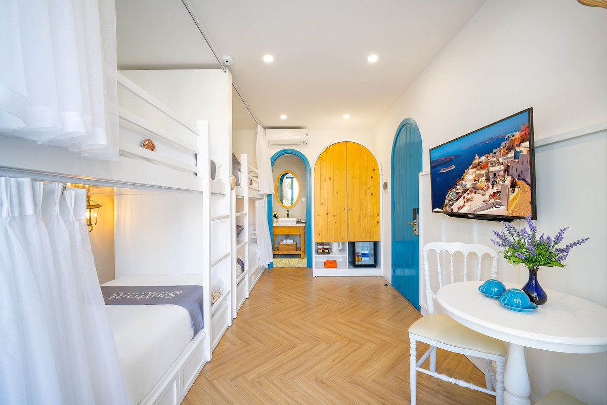 Quarto de hotel com beliches, banheiro pequeno, área de refeições com mesa e cadeiras, e uma janela grande com vista para uma cidade costeira.