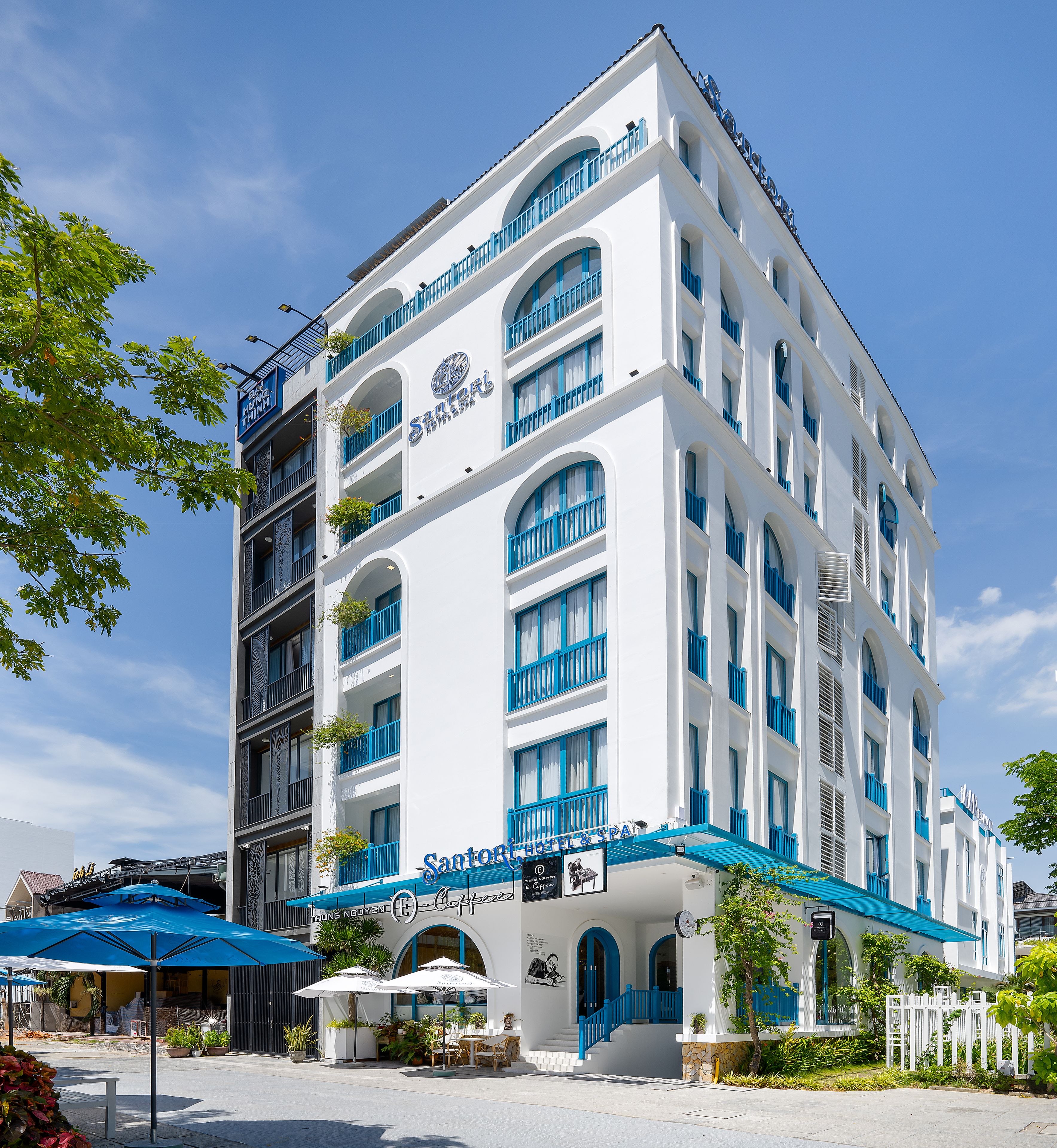 A multi-story hotel with a white facade and blue accents, featuring balconies and a coffee shop on the ground level.