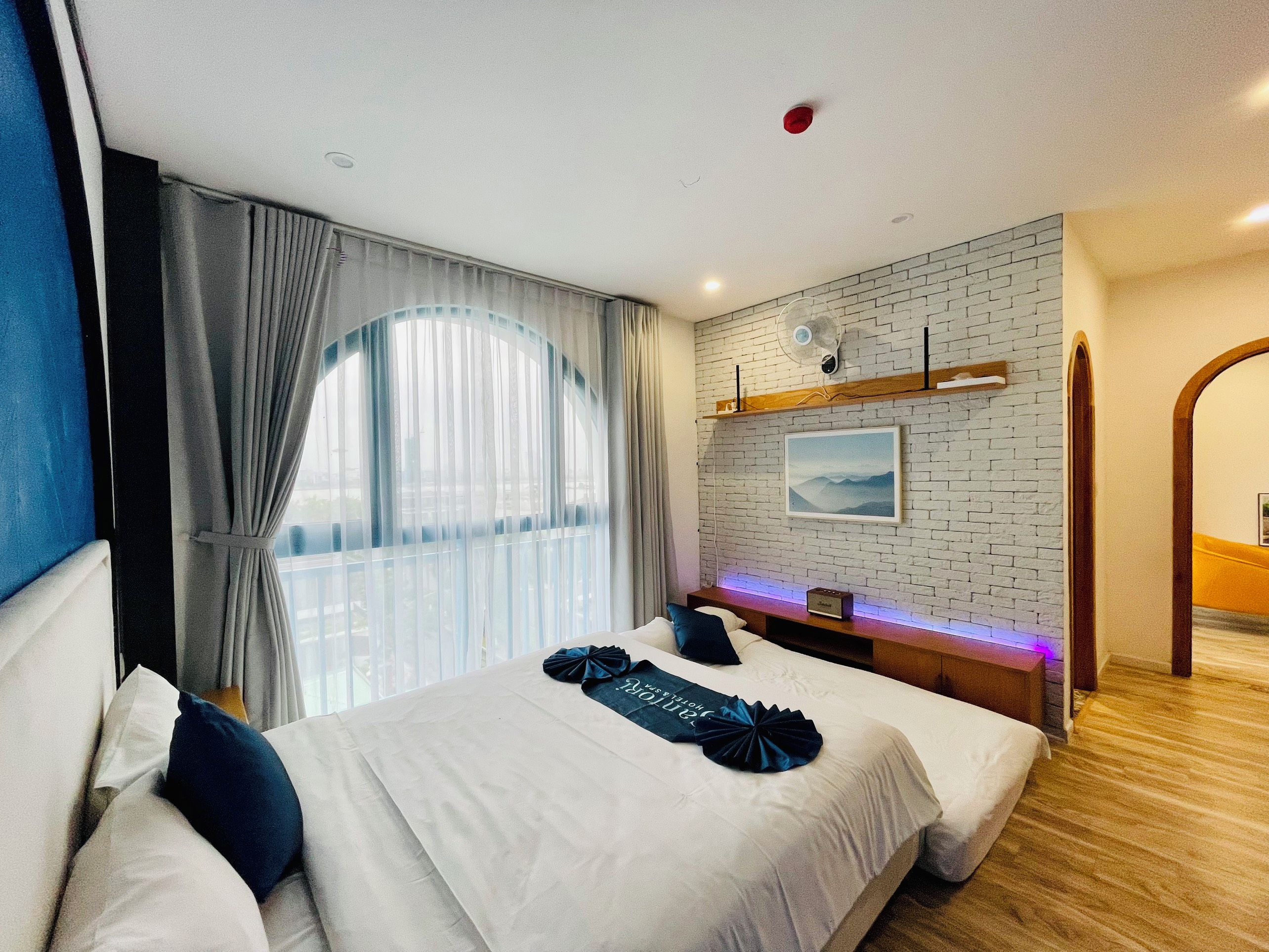 A hotel room with a large bed, a bedside table, a wall-mounted fan, and a window with curtains.