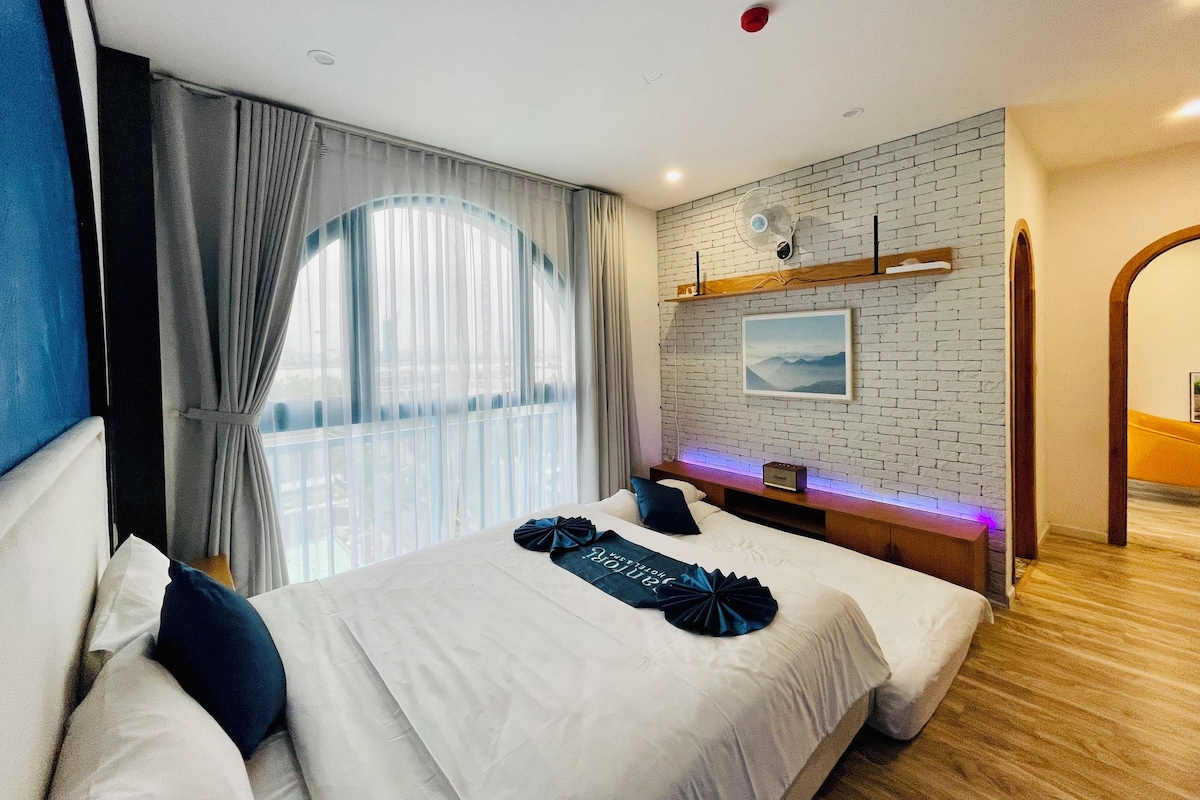Quarto de hotel com cama grande, mesa de cabeceira, ventoinha na parede e janela com cortinas.