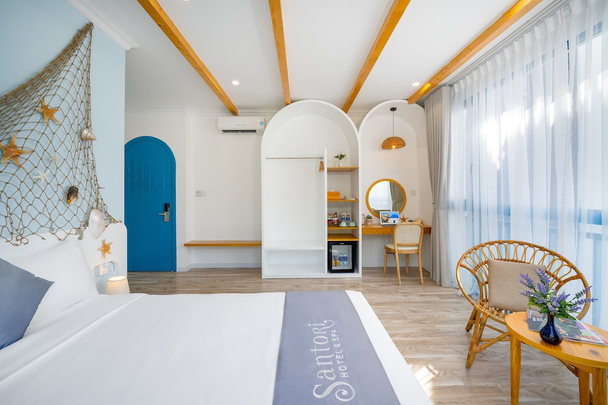 Quarto de hotel com cama, escrivaninha, cadeira, pequeno frigorífico e janela com cortinas.