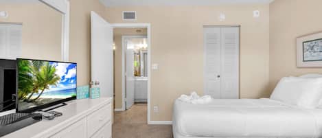 A hotel room with a bed, a desk with a computer, a television displaying a beach scene, and a door leading to a bathroom.